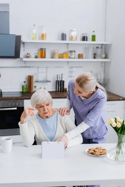 Junge Krankenschwester zeigt auf Kalender in der Nähe einer älteren Frau, die an Gedächtnisverlust leidet — Stockfoto