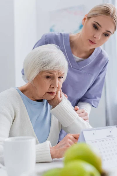 Junge Sozialarbeiterin in der Nähe einer betagten Frau blickt auf Kalender im verschwommenen Vordergrund — Stockfoto