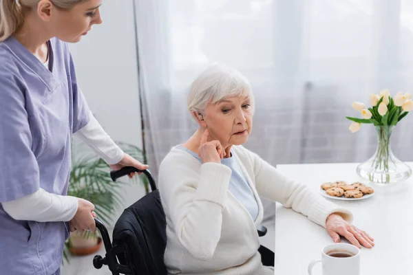 Donna anziana disabile che indica l'apparecchio acustico mentre è seduta sulla sedia a rotelle vicino all'assistente sociale — Foto stock