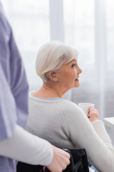 Disabled senior woman with hearing aid holding cup of tea near social worker on blurred foreground — Stock Photo