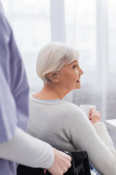 Donna anziana disabile con apparecchio acustico che tiene una tazza di tè vicino all'assistente sociale in primo piano sfocato — Foto stock