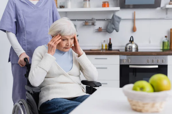 Aged disabled woman touching head while suffering from headache near social worker — Stock Photo