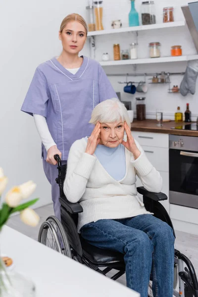 Jeune infirmière regardant la caméra près de femme âgée handicapée malade de démence — Photo de stock