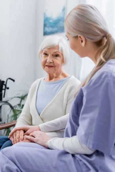 Enfermera joven tocando las manos de una anciana sentada en casa - foto de stock