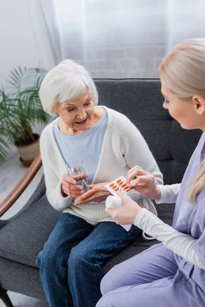 Krankenschwester gibt Seniorin, die mit Glas Wasser auf Couch sitzt, Medikamente — Stockfoto