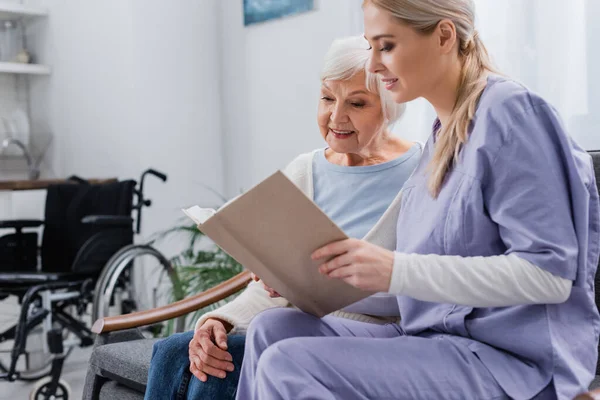 Junge Sozialarbeiterin liest Seniorin Buch, während sie auf Sofa sitzt — Stockfoto