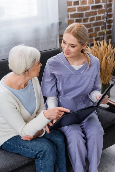 Aged woman pointing with finger at photo album while sitting on sofa with social worker — Stock Photo