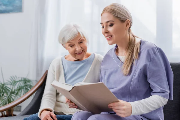 Lächelnde Krankenschwester liest glücklicher Seniorin auf der heimischen Couch Buch vor — Stockfoto