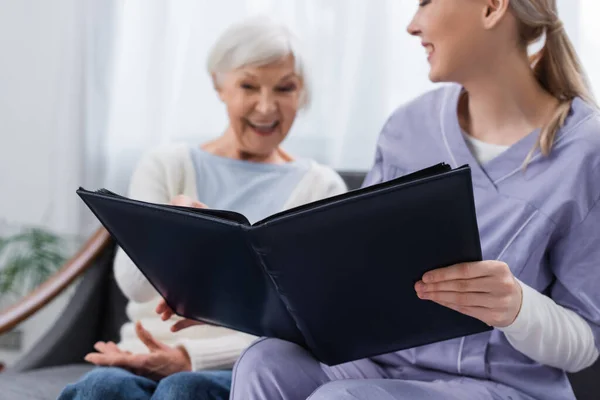 Fröhliche Seniorin mit Sozialarbeiterin schaut zu Hause auf Fotoalbum, verschwommener Hintergrund — Stockfoto