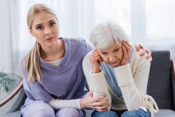 Young nurse looking at camera while embracing senior woman sitting with bowed head — Stock Photo