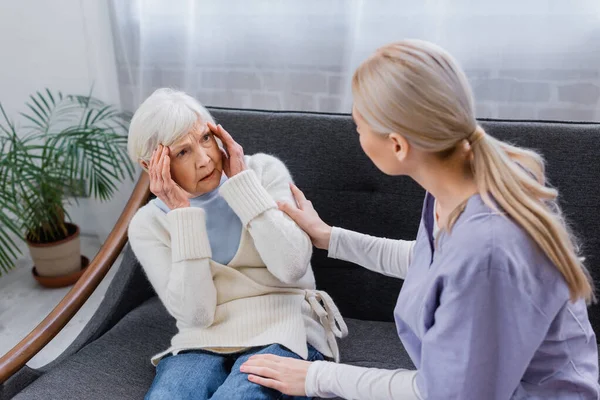 Aged woman, suffering from memory loss, looking at social worker touching her on sofa — Stock Photo