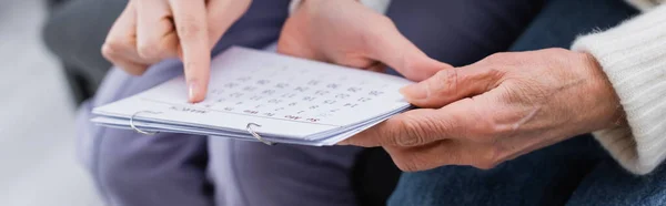 Vista recortada de la trabajadora social señalando el calendario cerca de la mujer anciana que sufre de pérdida de memoria, pancarta - foto de stock