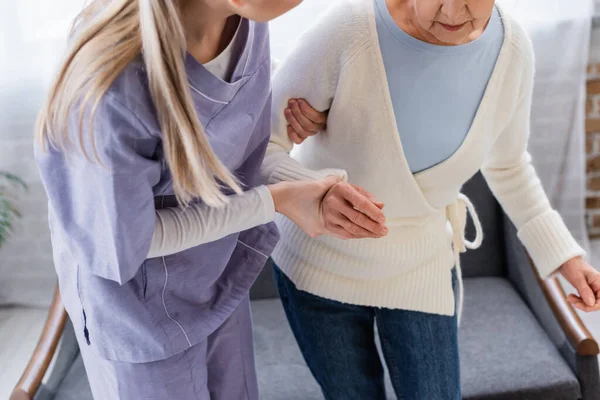 Partial view of social worker helping elderly woman getting up from sofa at home — Stock Photo