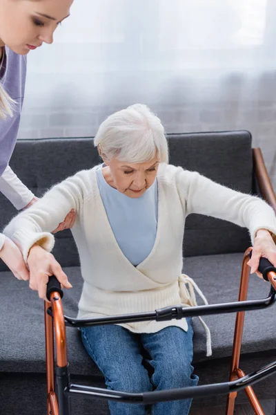 Social worker helping elderly woman getting up from sofa near medical walkers — Stock Photo