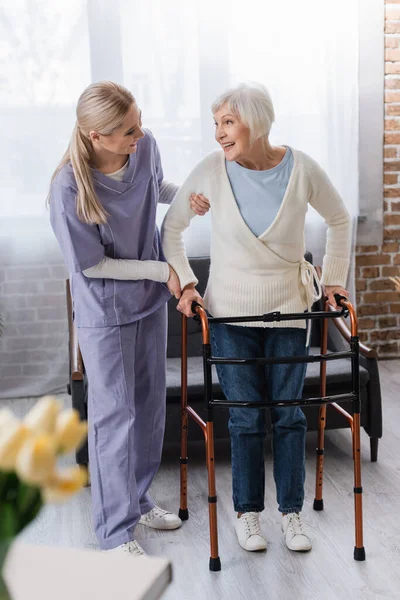 Young nurse supporting smiling senior woman walking in living room with medical walkers — Stock Photo