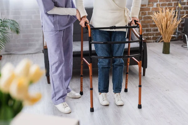 Visão parcial da enfermeira que assiste a mulher idosa durante a reabilitação com caminhantes médicos em casa — Fotografia de Stock