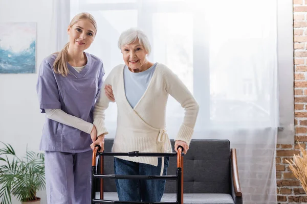 Enfermera feliz y mujer mayor mirando a la cámara cerca de los caminantes médicos en la sala de estar - foto de stock