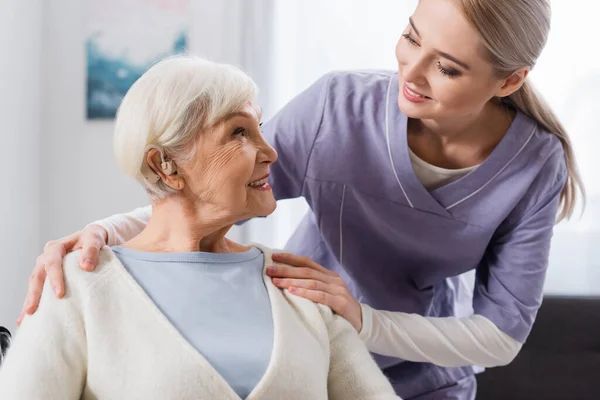 Sorridente assistente social abraçando ombro de mulher idosa com aparelho auditivo — Fotografia de Stock