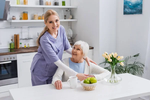 Junge Krankenschwester lächelt in die Kamera, während sie eine glückliche alte Frau in der Küche umarmt — Stockfoto