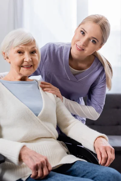 Infermiera felice e anziana donna handicappata sorridente mentre guarda la fotocamera — Foto stock