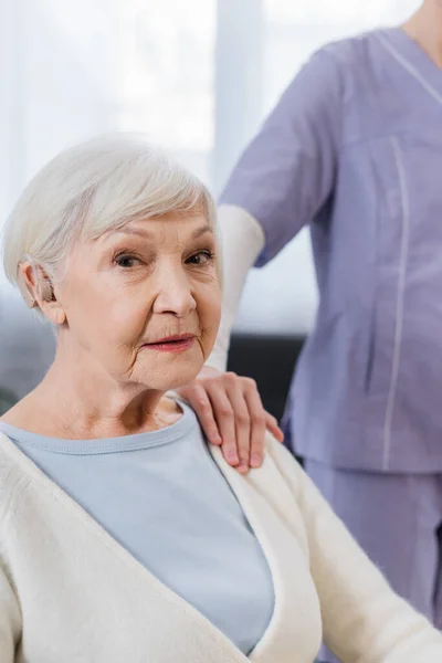 Alte Frau mit Hörgerät blickt in Kamera in der Nähe von Sozialarbeiterin, die ihre Schulter berührt — Stockfoto