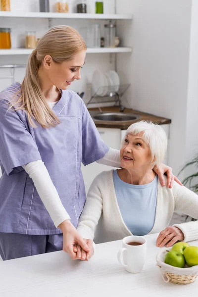 Jovem enfermeira segurando a mão e abraçando o ombro da mulher idosa alegre na cozinha — Fotografia de Stock