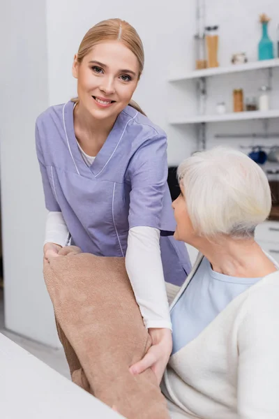 Happy social worker looking at camera while giving soft blanket to elderly woman — Stock Photo