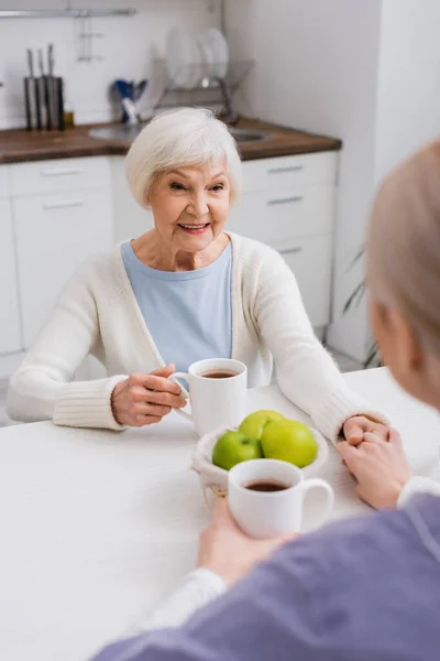 Happy elderly woman holding hands with social worker near cups of tea in kitchen, blurred foreground — Stock Photo