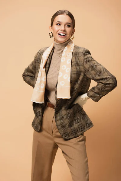 Femme gaie dans des vêtements élégants debout avec les mains sur les hanches et regardant loin sur beige — Photo de stock