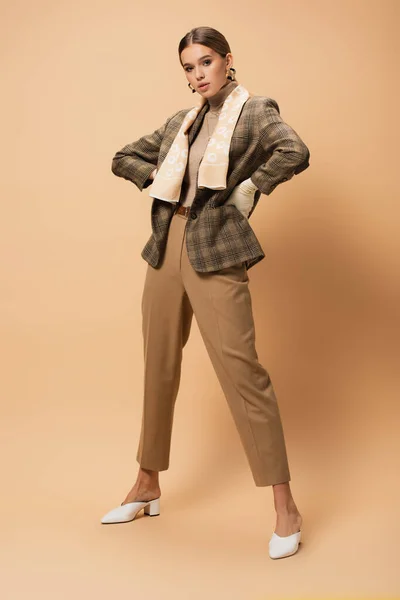 Vue pleine longueur de la femme élégante en blazer et pantalon posant avec les mains sur les hanches sur beige — Photo de stock