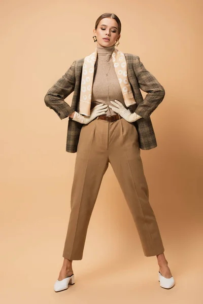 Elegante mujer en chaqueta marrón y pantalones de pie con las manos en la cintura sobre fondo beige - foto de stock