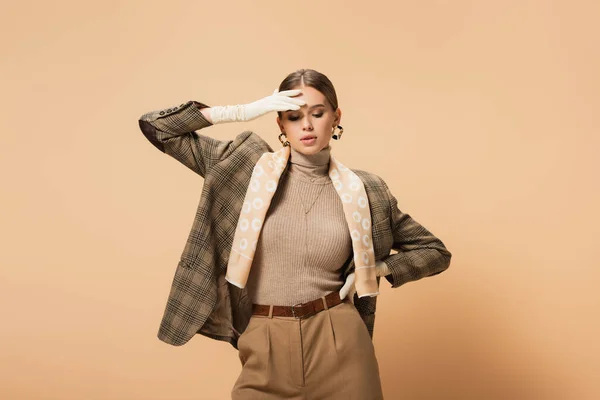 Femme à la mode en blazer et pantalon touchant le front tout en posant avec la main sur la hanche sur beige — Photo de stock