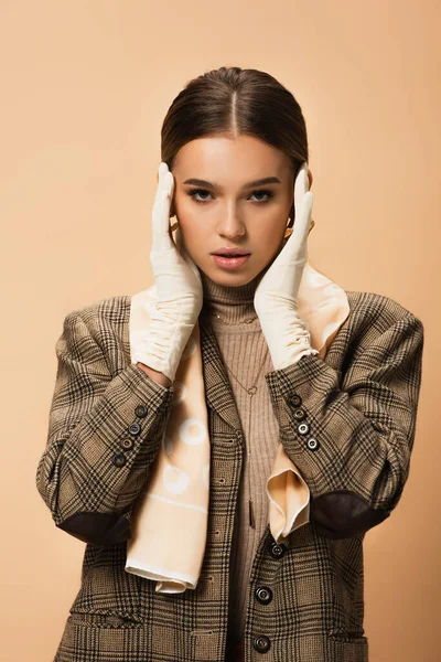 Mujer joven en blazer de moda y guantes blancos posando con las manos cerca de la cara aislada en beige - foto de stock