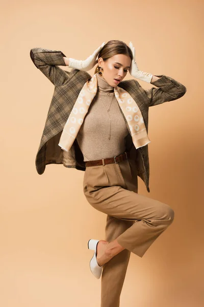 Trendy woman in blazer and trousers holding hands near head while standing on one leg on beige — Stock Photo