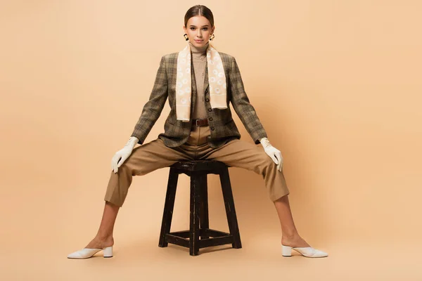 Mujer joven en blazer de moda, pantalones, zapatos blancos y guantes sentados en taburete de madera en beige - foto de stock