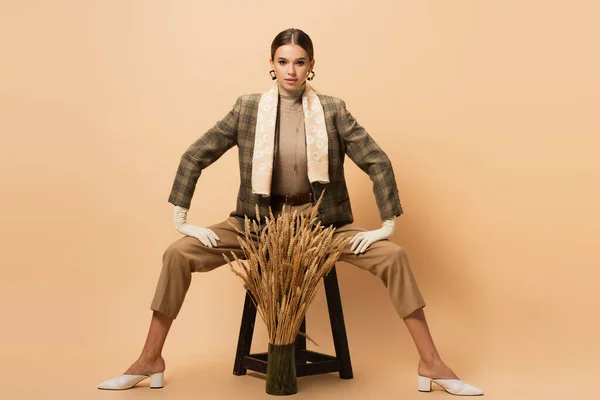 Stylish woman in trousers and blazed sitting on wooden stool near spikelets on beige — Stock Photo