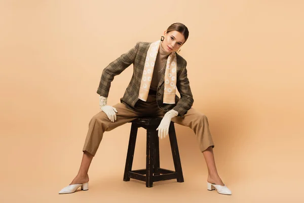 Stylish woman in blazer and pants looking at camera while sitting on wooden stool on beige — Stock Photo
