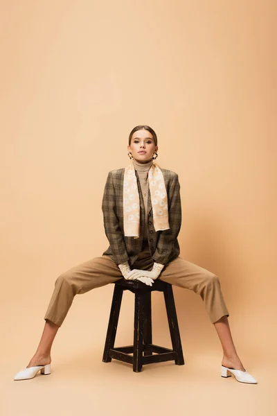 Вид спереди стильной женщины в коричневом блейзере и брюках, позирующей на деревянной табуретке бежевого цвета — стоковое фото