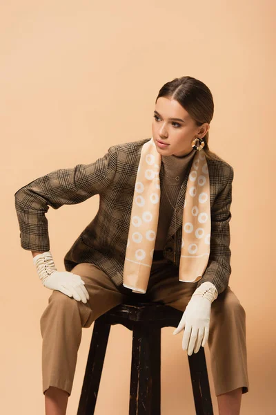 Fashionable woman in blazer, pants and gloves looking away while sitting on stool isolated on beige — Stock Photo