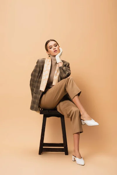 Mujer de moda en chaqueta y pantalones sentados en taburete de madera sobre fondo beige - foto de stock