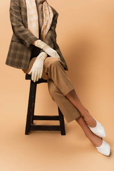 Обрезанный вид женщины в штанах, клетчатый пиджак, белые перчатки и обувь, сидящие на бежевом — стоковое фото