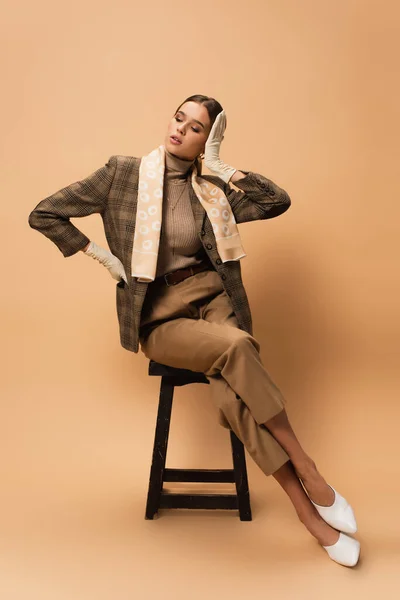 Fashionable woman in brown blazer and trousers touching hair while sitting on beige — Stock Photo