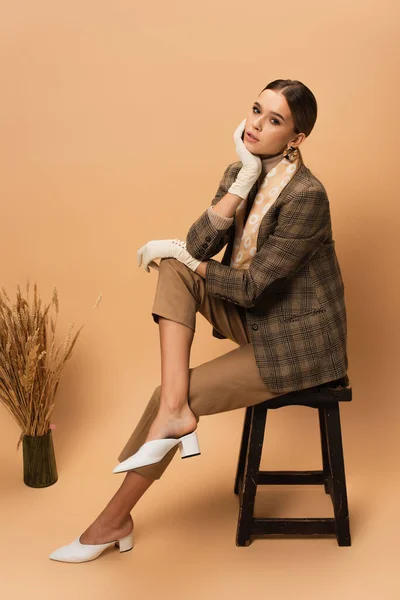 Elegante donna in blazer a quadretti, pantaloni, scarpe bianche e guanti seduta vicino al vaso con spillette su beige — Foto stock