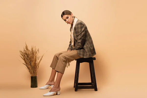 Jeune femme en blazer tendance et pantalon assis sur tabouret près du vase avec des épillets sur beige — Photo de stock