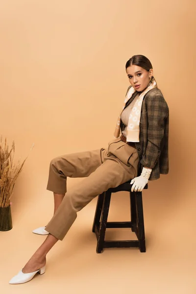 Полный вид стильной женщины в брюках и клетчатом пиджаке на деревянном стуле на бежевом фоне — стоковое фото