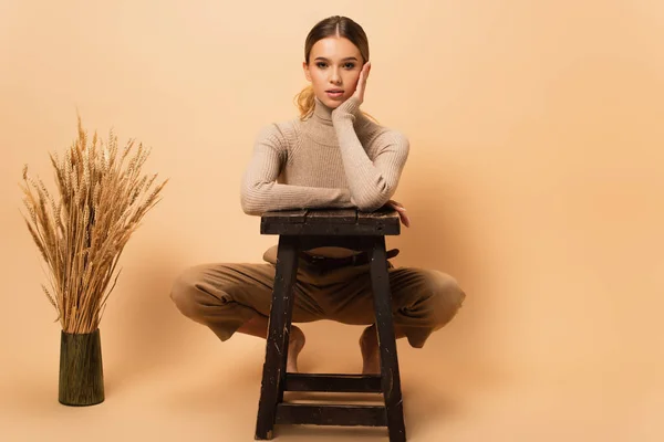 Fashionable barefoot woman posing near stool and vase with spikelets on beige background — Stock Photo