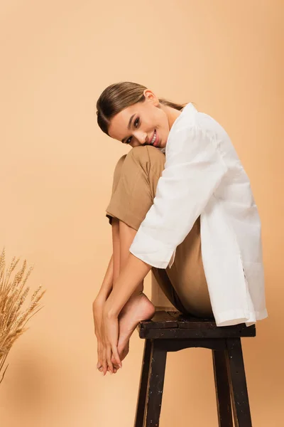 Lächelnde Frau in stilvoller Kleidung, die barfuß auf einem Stuhl sitzt und in beige in die Kamera schaut — Stockfoto