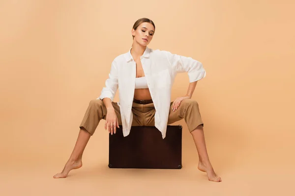 Mujer descalza con estilo en pantalones y camisa blanca posando en una maleta vintage sobre fondo beige - foto de stock