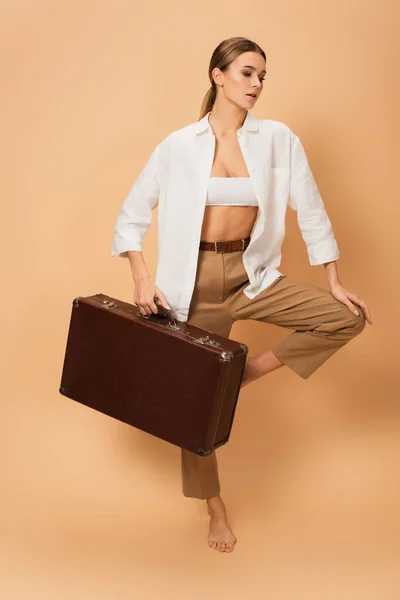 Trendy woman in trousers and white shirt standing on one leg with retro suitcase on beige background — Stock Photo