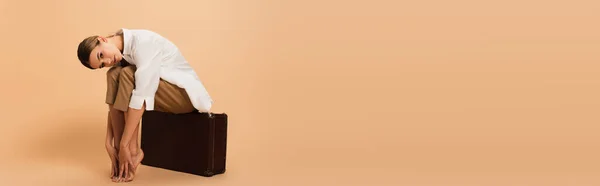 Femme pieds nus dans des vêtements élégants assis sur une valise vintage sur fond beige, bannière — Photo de stock